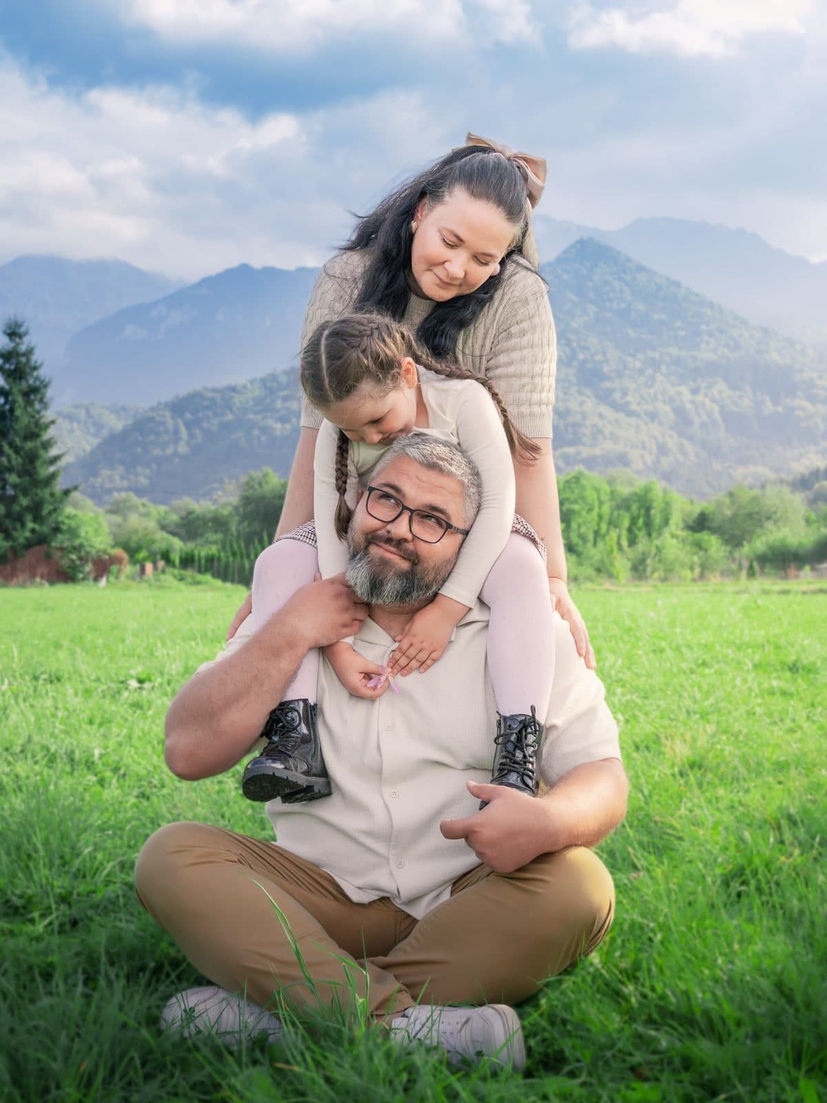 Fetița pe umerii tatălui, mama aplecată deasupra lor, pe o pajiște cu munți