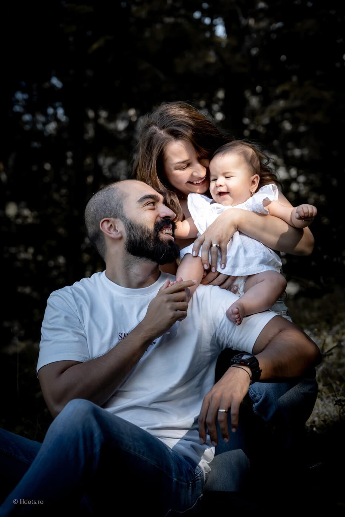 Familie tânără îmbrățișată în pădure, tatăl sărută bebelușul pe obraz
