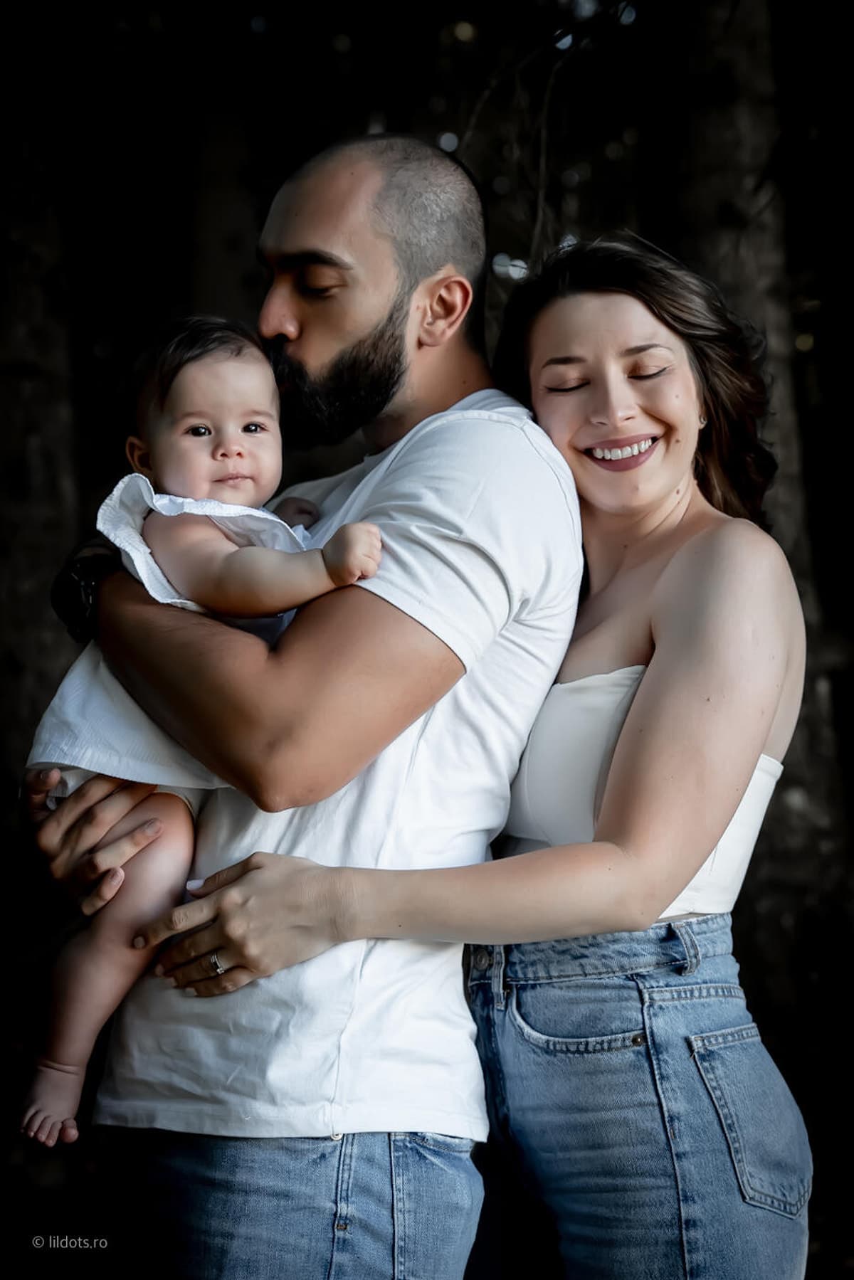 Tatăl o sărută pe bebelușă pe creștet, mama zâmbește cu ochii închiși lângă el